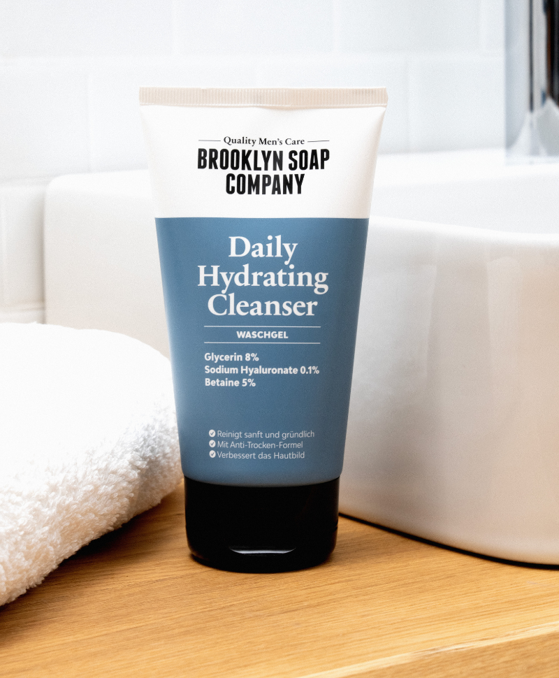 Brooklyn Soap Company Daily Hydrating Cleanser – ein blau-weißer Reinigungsgel-Tube auf einem Holzwaschtisch neben einem weißen Handtuch und einer weißen Badewanne im Hintergrund.