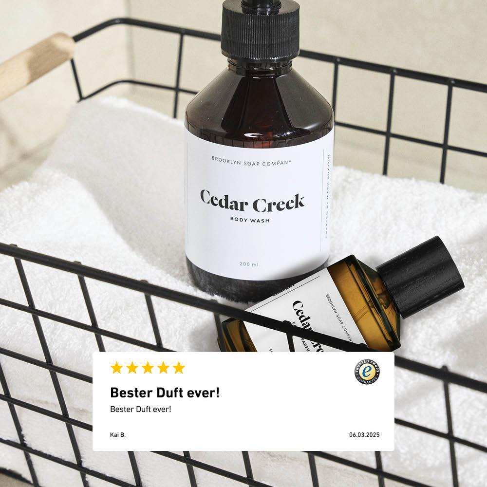 Zwei Produkte aus der Cedar Creek Serie von Brooklyn Soap Company – Body Wash und Eau de Parfum – stehen in einem schwarzen Drahtkorb auf einem weißen Handtuch. Im Vordergrund eine eingeblendete 5-Sterne-Bewertung mit dem Kommentar „Bester Duft ever!“.