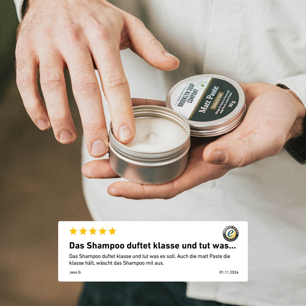 Ein Mann öffnet eine silberne Dose der Brooklyn Soap Company Matt Paste, während er mit dem Finger etwas Produkt entnimmt – darunter eine Kundenbewertung über Duft und Qualität des Shampoos.