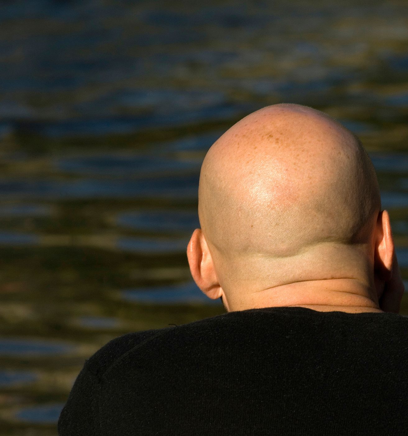 Eine Person mit glatt rasierter Kopfhaut ist von hinten vor einem hellen, neutralen Hintergrund zu sehen.